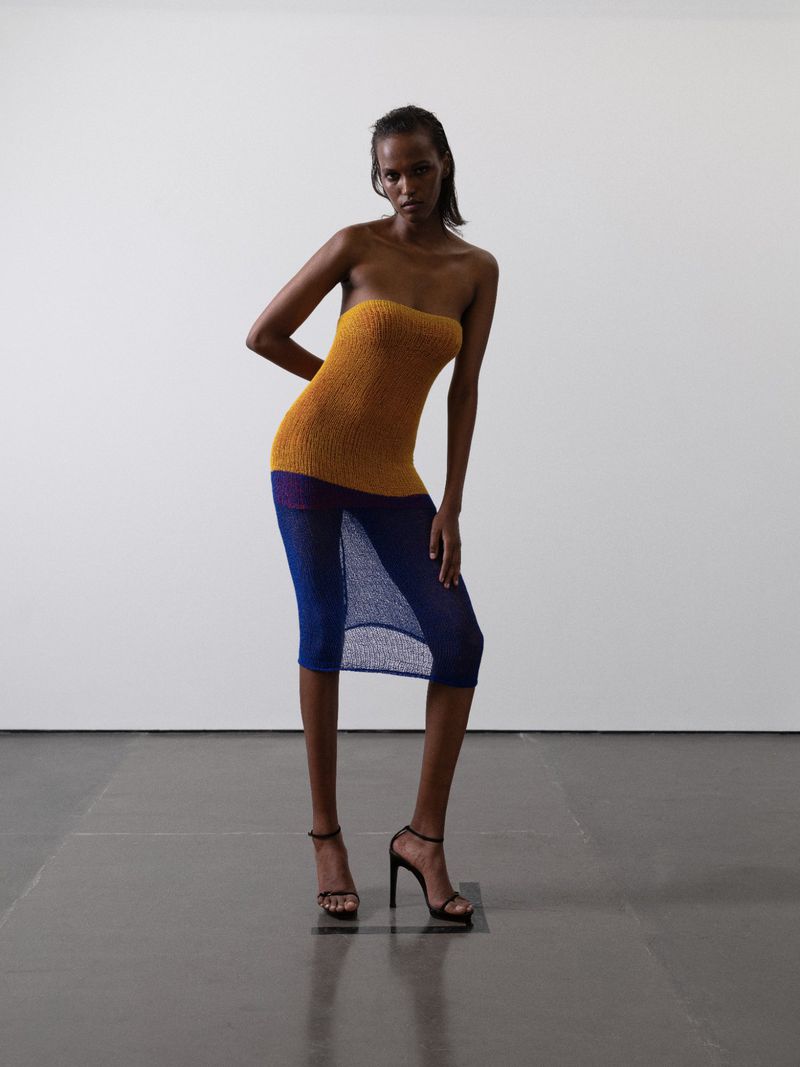 A slender, dark-skinned woman models a sleeveless knit dress with heels in a photo studio with a neutral background, showcasing fashion.