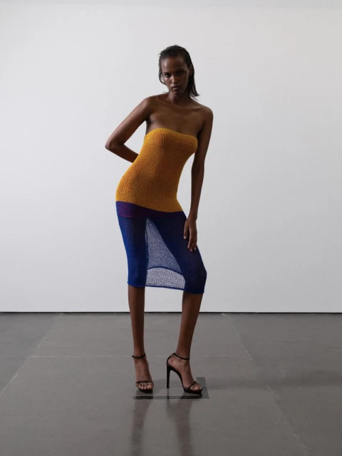 A slender, dark-skinned woman models a sleeveless knit dress with heels in a photo studio with a neutral background, showcasing fashion.