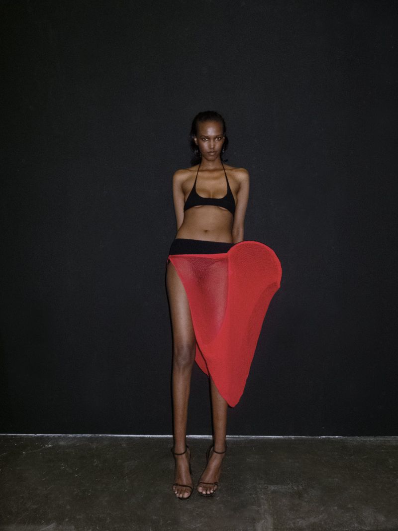 Woman in bikini and red skirt poses against black backdrop, showcasing fashion and style with studio lighting and a confident expression.