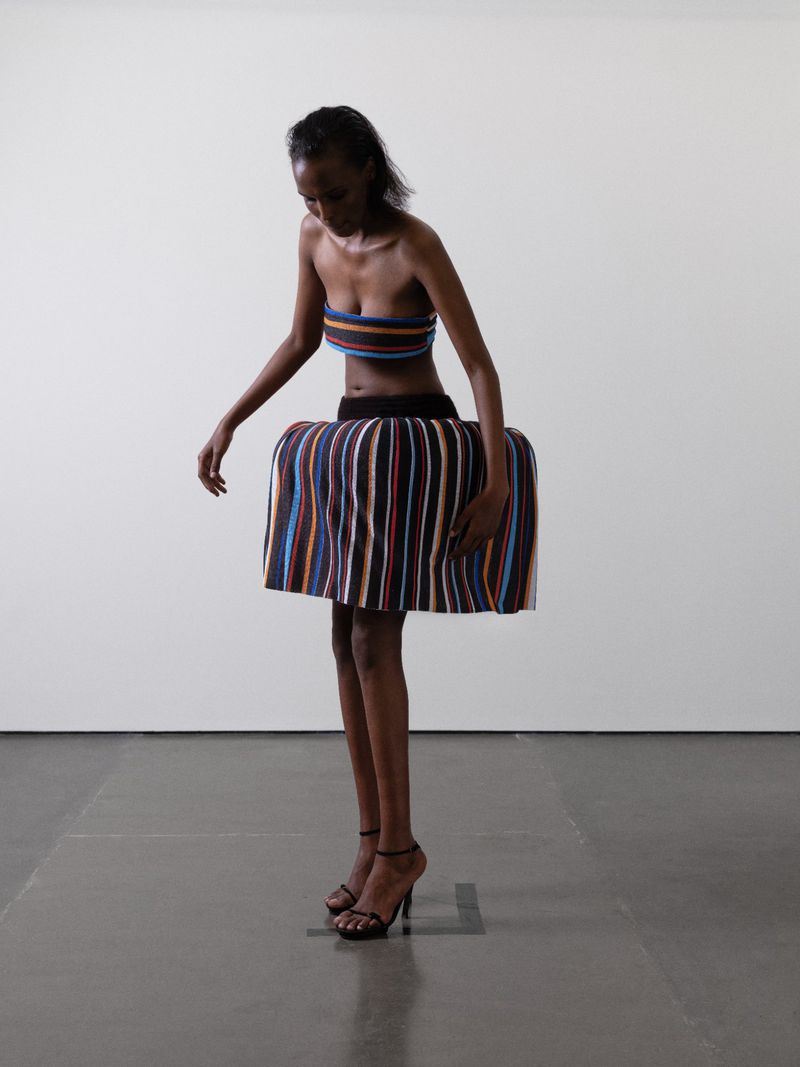 Elegant studio shot of a slender Black woman in a striped tube top and sculptural skirt, accessorized with strappy heeled shoes, against a white backdrop.