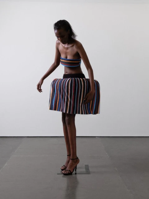 Elegant studio shot of a slender Black woman in a striped tube top and sculptural skirt, accessorized with strappy heeled shoes, against a white backdrop.
