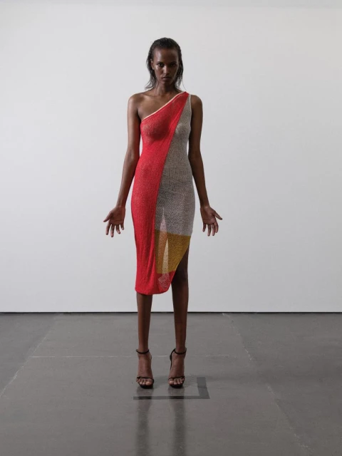 A slender woman in a red, silver, and gold asymmetric dress and high heels stands in a bright minimalist studio, fashion forward.