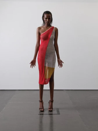 A slender woman in a red, silver, and gold asymmetric dress and high heels stands in a bright minimalist studio, fashion forward.