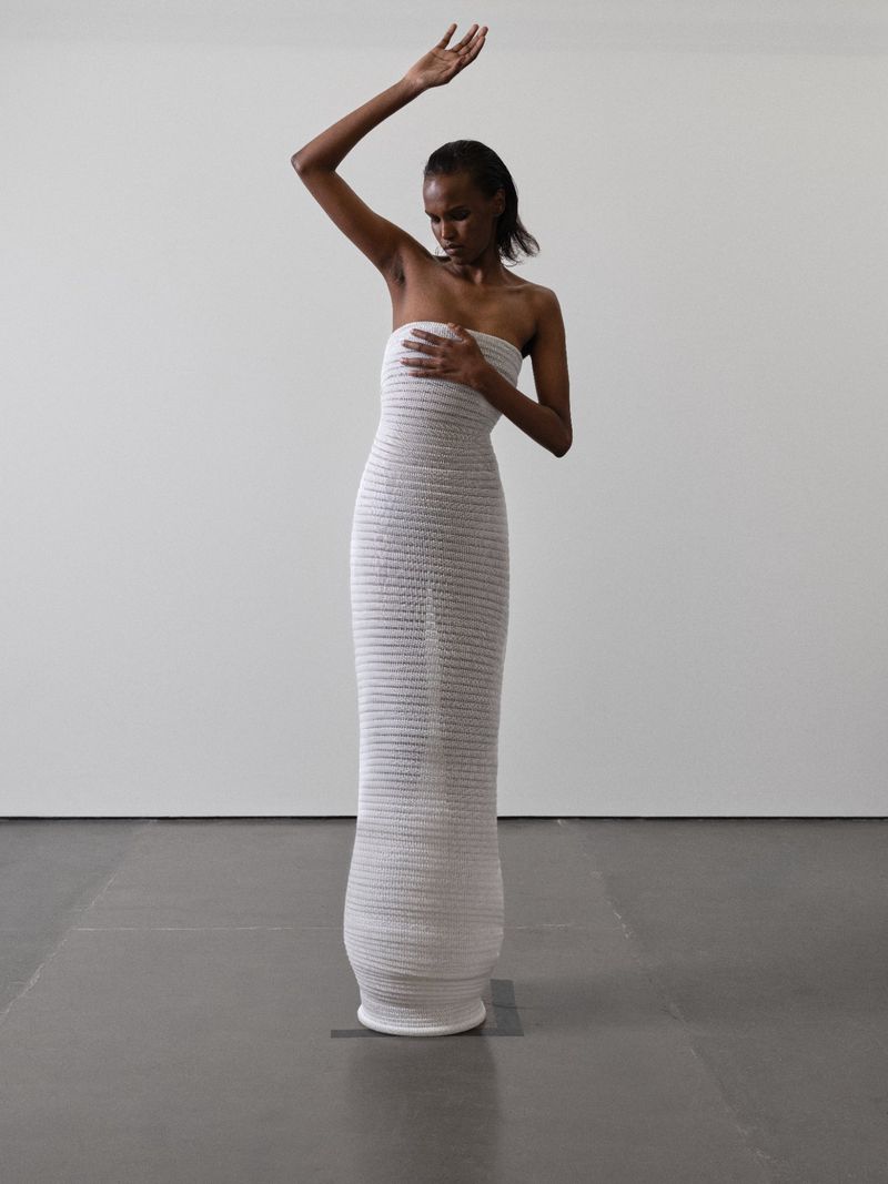 An African-American adult female fashion model poses in a floor-length, textured, strapless white dress with one arm raised against a white wall.