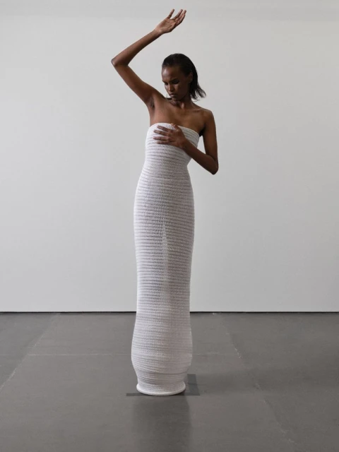 An African-American adult female fashion model poses in a floor-length, textured, strapless white dress with one arm raised against a white wall.