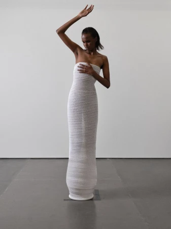 An African-American adult female fashion model poses in a floor-length, textured, strapless white dress with one arm raised against a white wall.