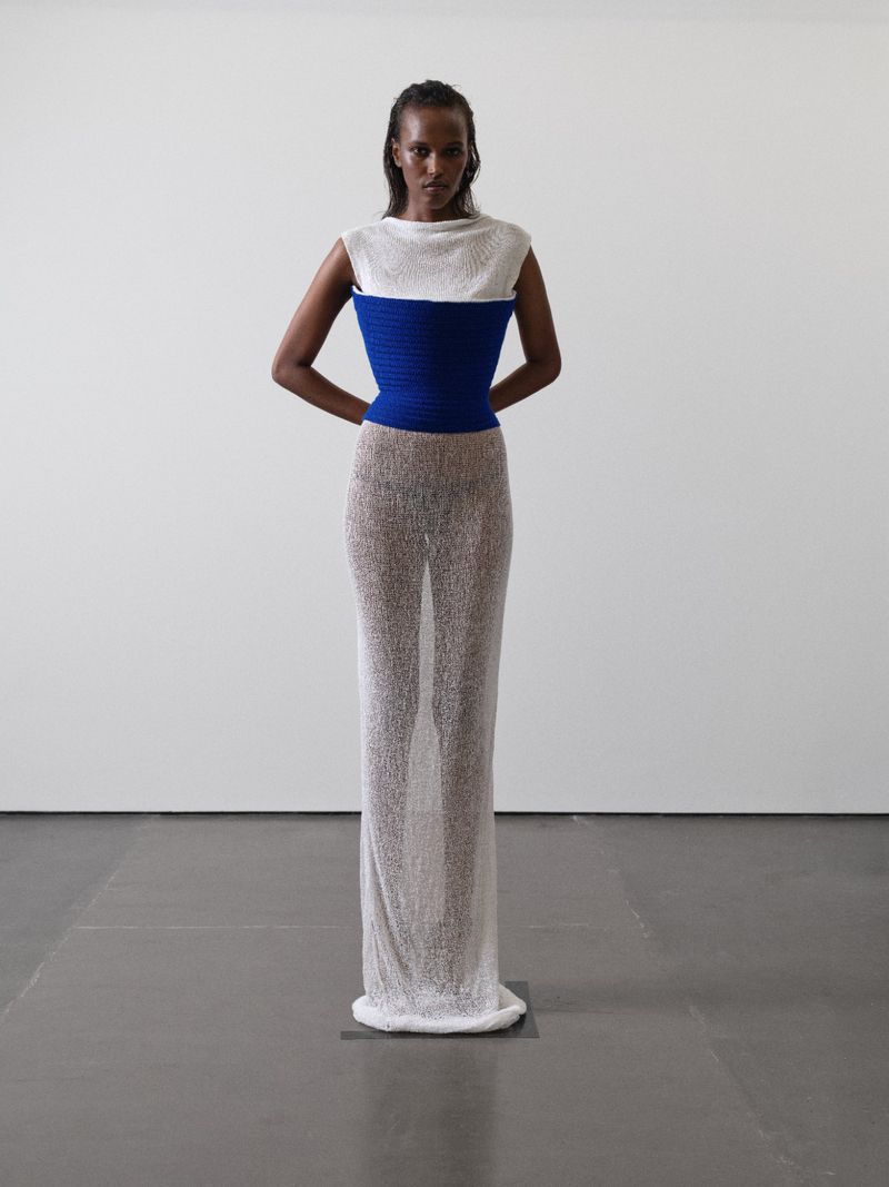 A woman stands in a photo studio wearing a white and blue top with a semi-sheer dress. The image has studio lighting and a white background.