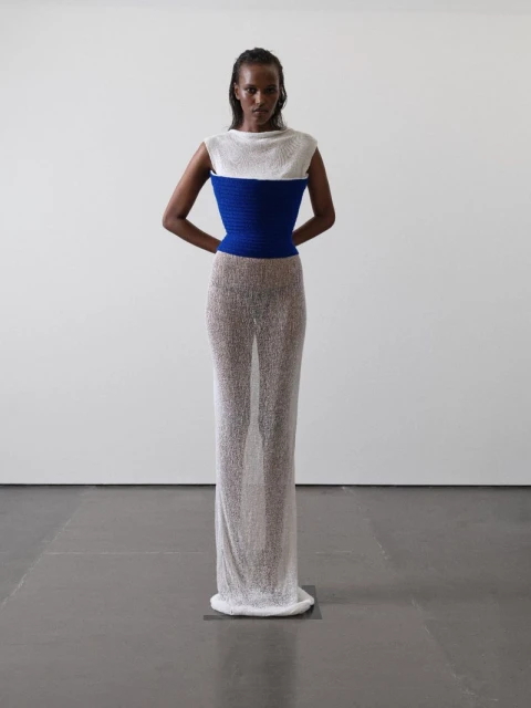 A woman stands in a photo studio wearing a white and blue top with a semi-sheer dress. The image has studio lighting and a white background.