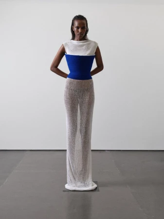 A woman stands in a photo studio wearing a white and blue top with a semi-sheer dress. The image has studio lighting and a white background.