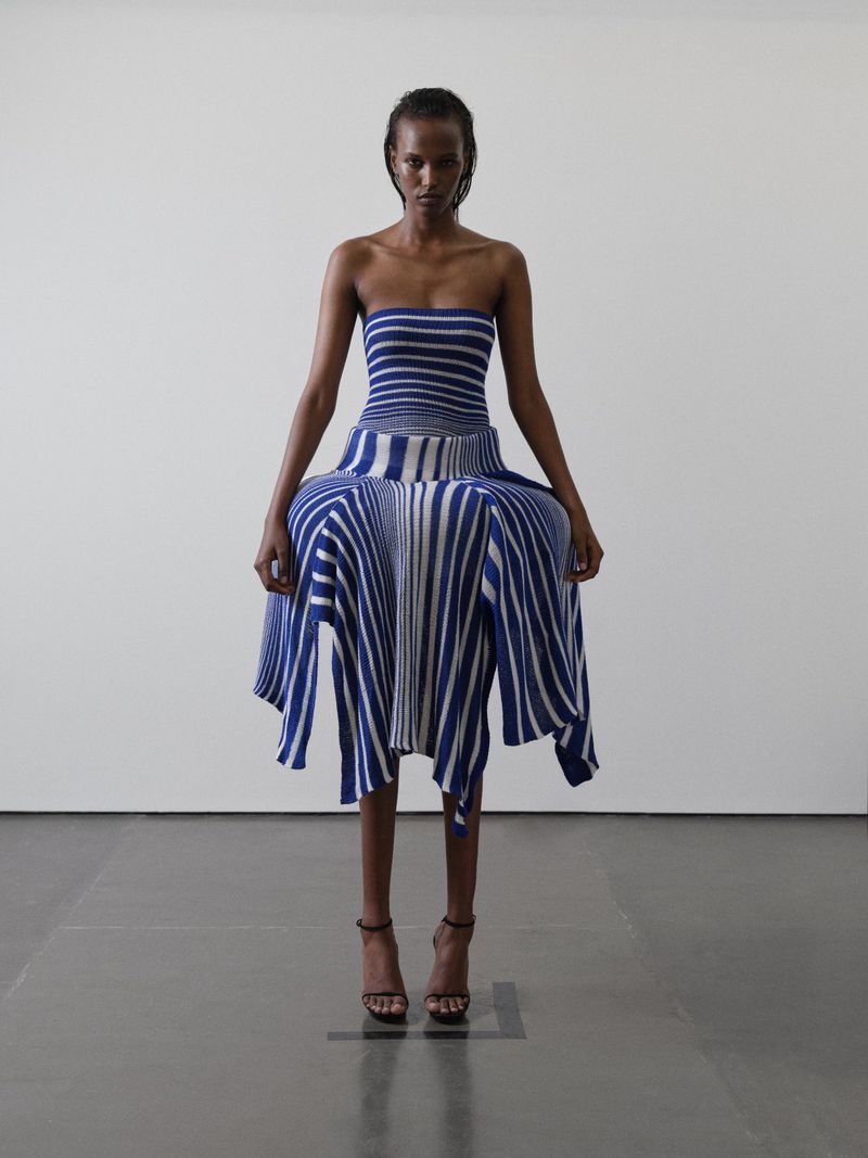 An adult female poses in a blue and white striped strapless dress and heeled sandals in a studio setting against a white wall.