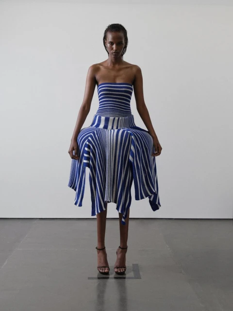 An adult female poses in a blue and white striped strapless dress and heeled sandals in a studio setting against a white wall.