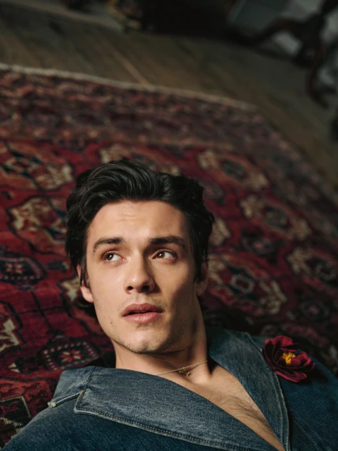 Portrait of a young adult male with dark hair lying on an ornate rug, wearing a denim jacket and a flower accessory.