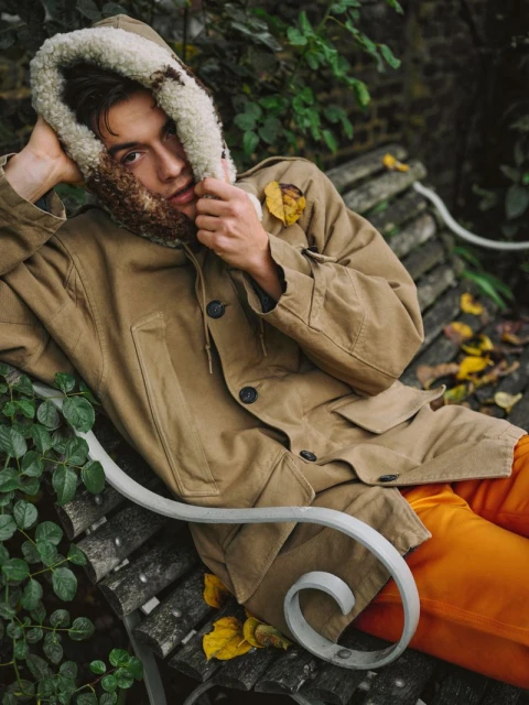 Adult male in tan hooded jacket, bright orange pants, and patterned scarf, reclines on a park bench with leaves and lush greenery.