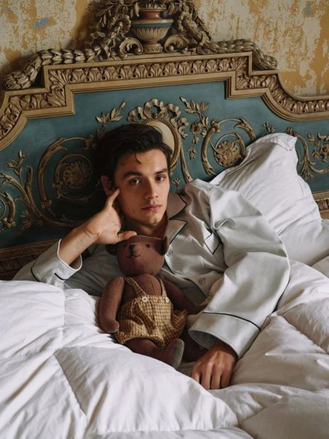 Adult male in white pajamas lying in bed with a teddy bear against an ornate headboard in a home interior with neutral colors.