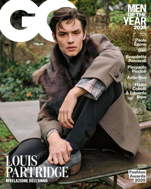 GQ Magazine cover featuring Louis Partridge in a beige overcoat with a fur stole, sitting outdoors on a metal bench with greenery behind him.
