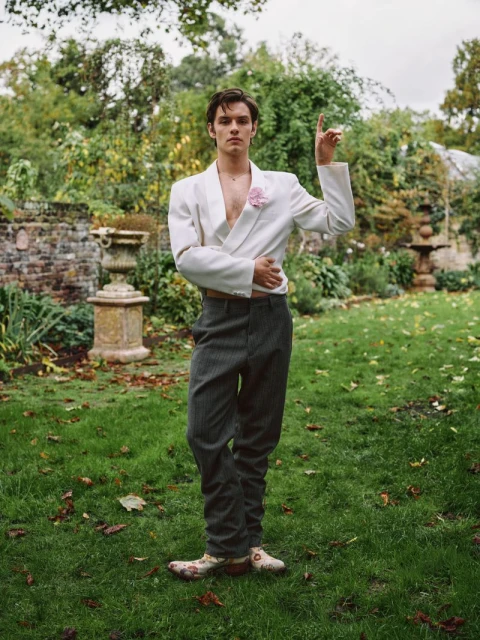 Full shot of a male model in a white suit jacket and grey pants posing in a garden with a variety of green plants.