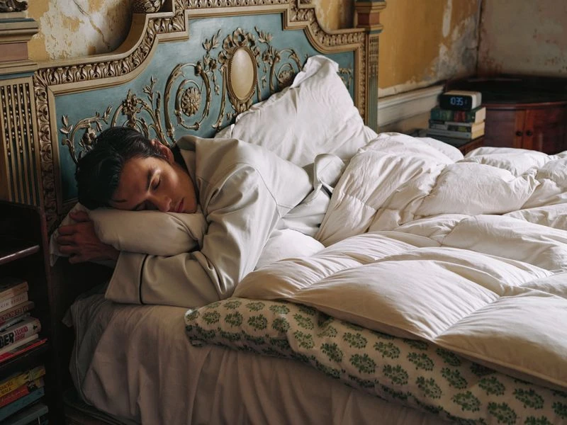 Adult man sleeping in a cozy bed with white linen bedding, ornate headboard, and a bedside table in a warmly lit interior.