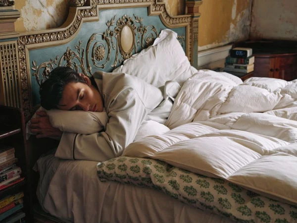 Adult man sleeping in a cozy bed with white linen bedding, ornate headboard, and a bedside table in a warmly lit interior.