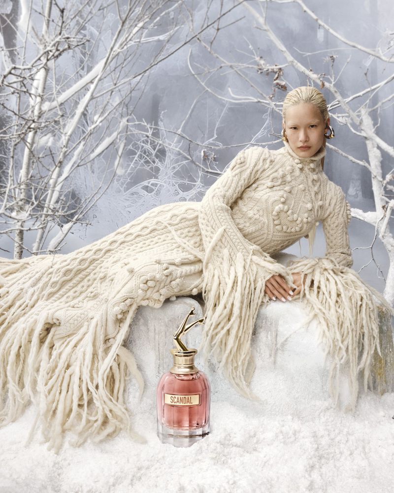 Fashion image: Asian woman poses in a long, textured, white fringe dress outdoors with a bottle of Scandal perfume in snowy scene.