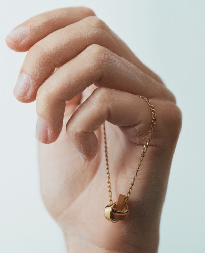 Close-up of an adult hand holding a gold necklace with a leather and gold ring pendant against a white background in portrait orientation.