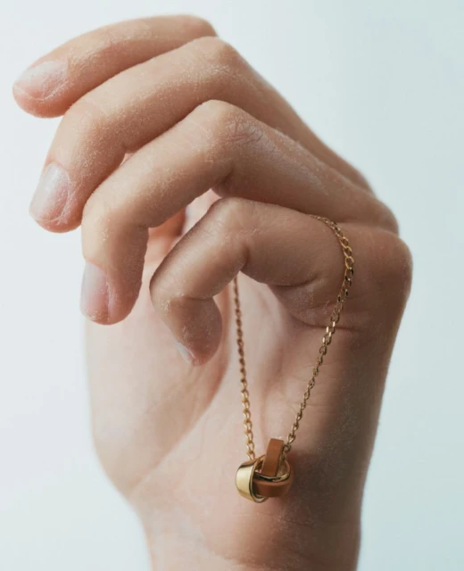 Close-up of an adult hand holding a gold necklace with a leather and gold ring pendant against a white background in portrait orientation.