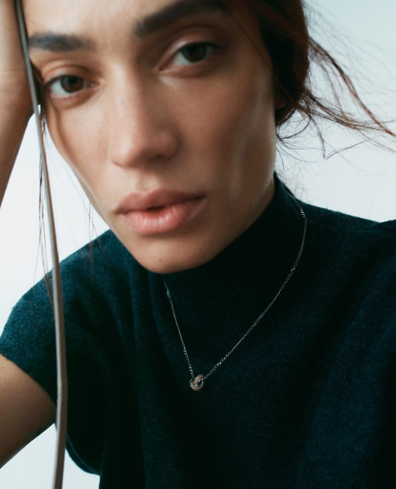 Close-up portrait of a woman wearing a dark green turtleneck and a silver pendant necklace against a white background