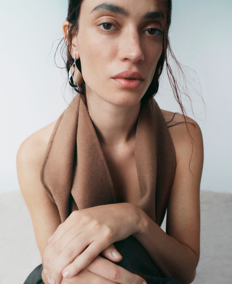 Portrait of an adult female model with tan scarf, silver earrings, dark hair, and light skin tone against a neutral grey background.