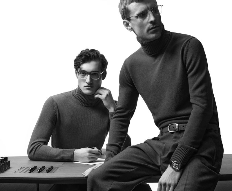 Two male models wearing glasses and turtlenecks sit at a table with pens and paper in a black and white indoor fashion shot.