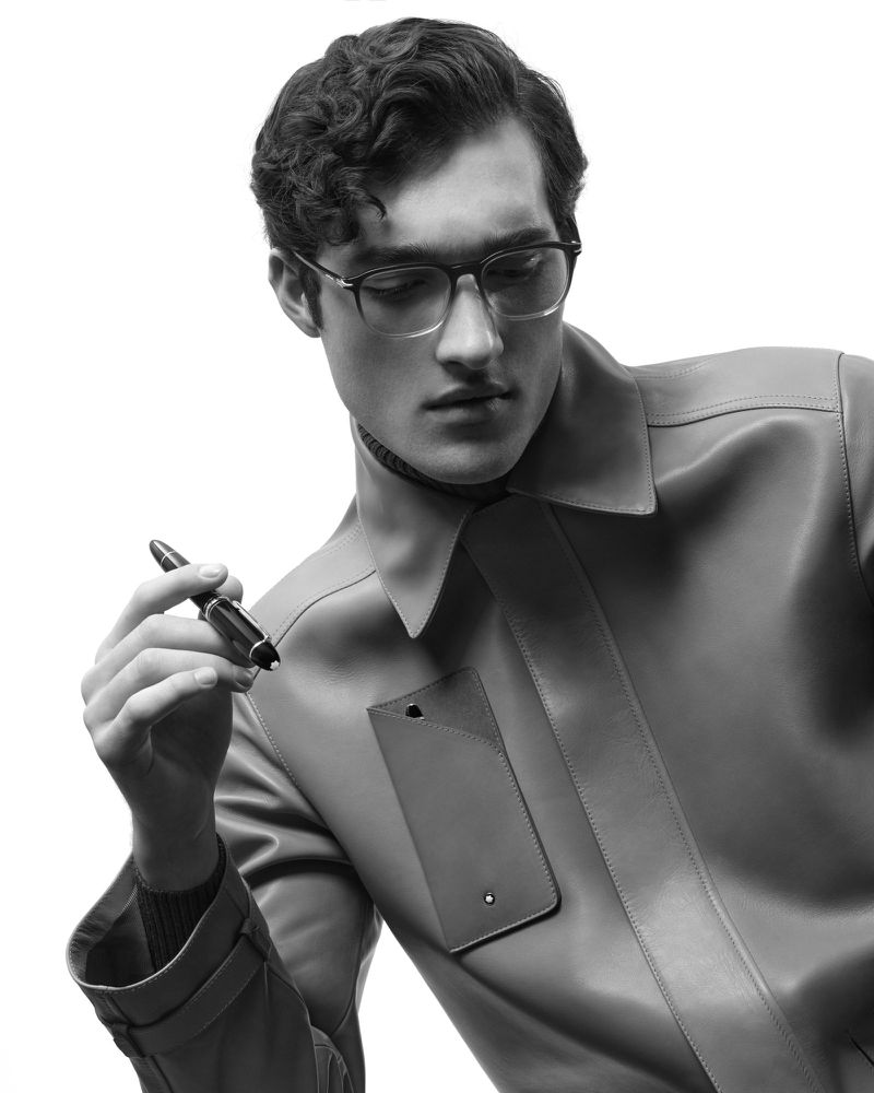 Studio shot of a male model wearing glasses and a leather jacket, holding a pen in a black-and-white image on a white background.
