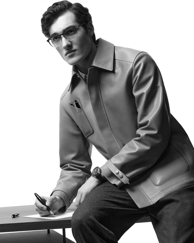 Monochrome portrait of a man in glasses and leather jacket writing on paper with a pen while sitting at a desk against a white background.