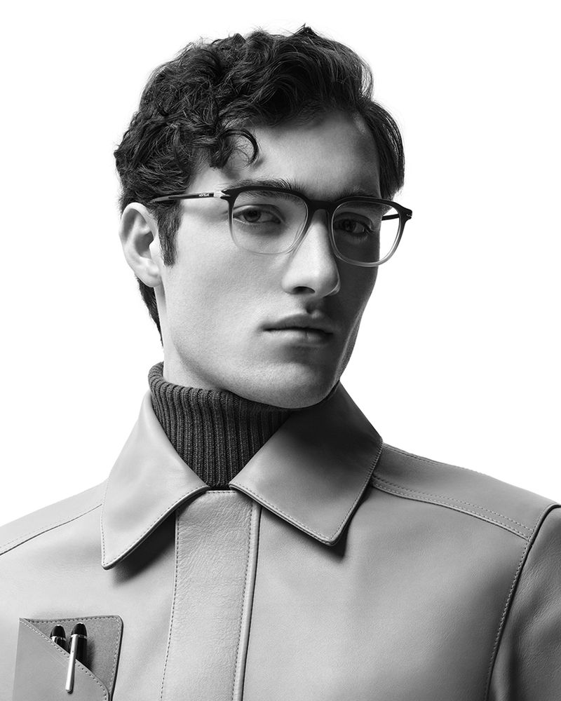A black and white portrait of a man with curly hair wearing glasses, a turtleneck sweater, and a coat against a bright white background.