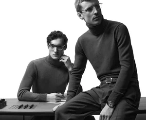 Two men wearing turtlenecks and eyeglasses posing at a dark desk against a plain white studio background.