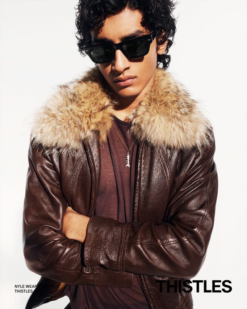 Portrait of a young man with sunglasses, wearing a brown leather jacket with a fur collar, and silver pendant against a white background.