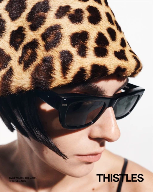 Close-up of a woman in sunglasses and a leopard-print hat against a white background, showcasing a fashion-forward editorial look.