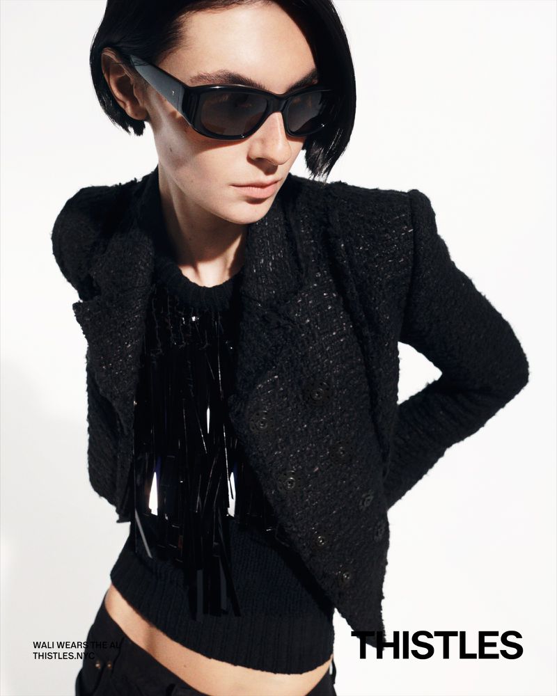 Woman in black jacket and sunglasses posing against a white backdrop. The shot emphasizes fashion and style in a studio environment.