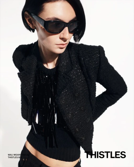Woman in black jacket and sunglasses posing against a white backdrop. The shot emphasizes fashion and style in a studio environment.