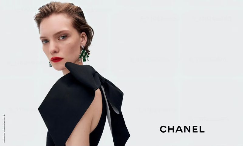 Fashion shot of a woman with short brown hair, red lipstick, and wearing green earrings and a black dress with a bow, on a white background.