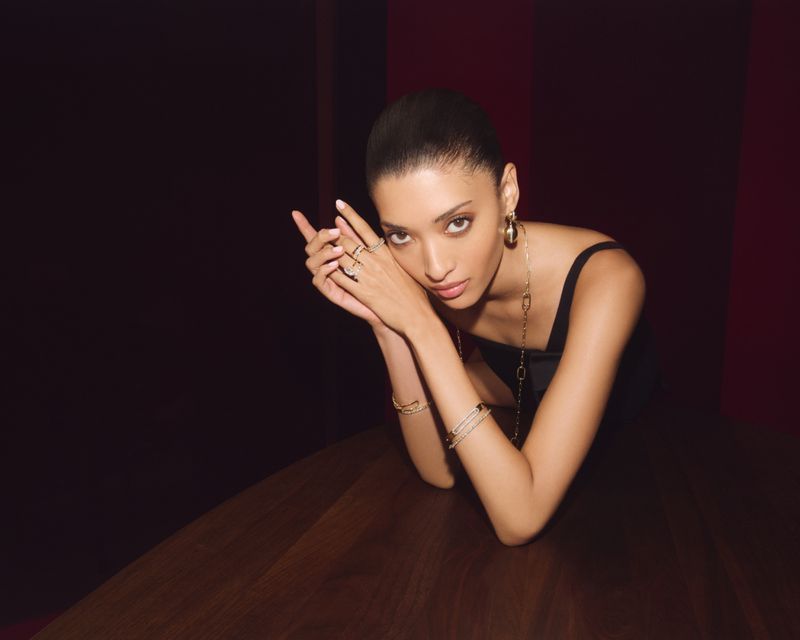 Portrait of a woman with dark hair pulled back, wearing jewelry and resting her chin on her clasped hands in front of a wooden table.