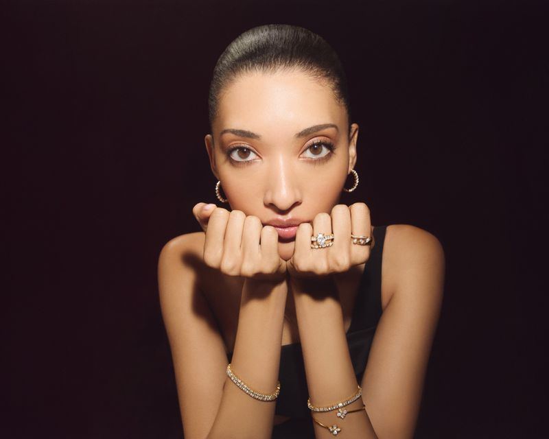 Model Aqua Parios resting chin on hands wearing diamond rings, bracelets, and hoop earrings against a dark backdrop.