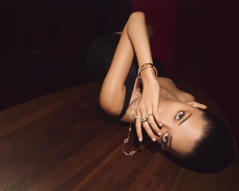 Model lying on a dark wooden surface wearing gold rings, gold bracelets, and a long gold necklace