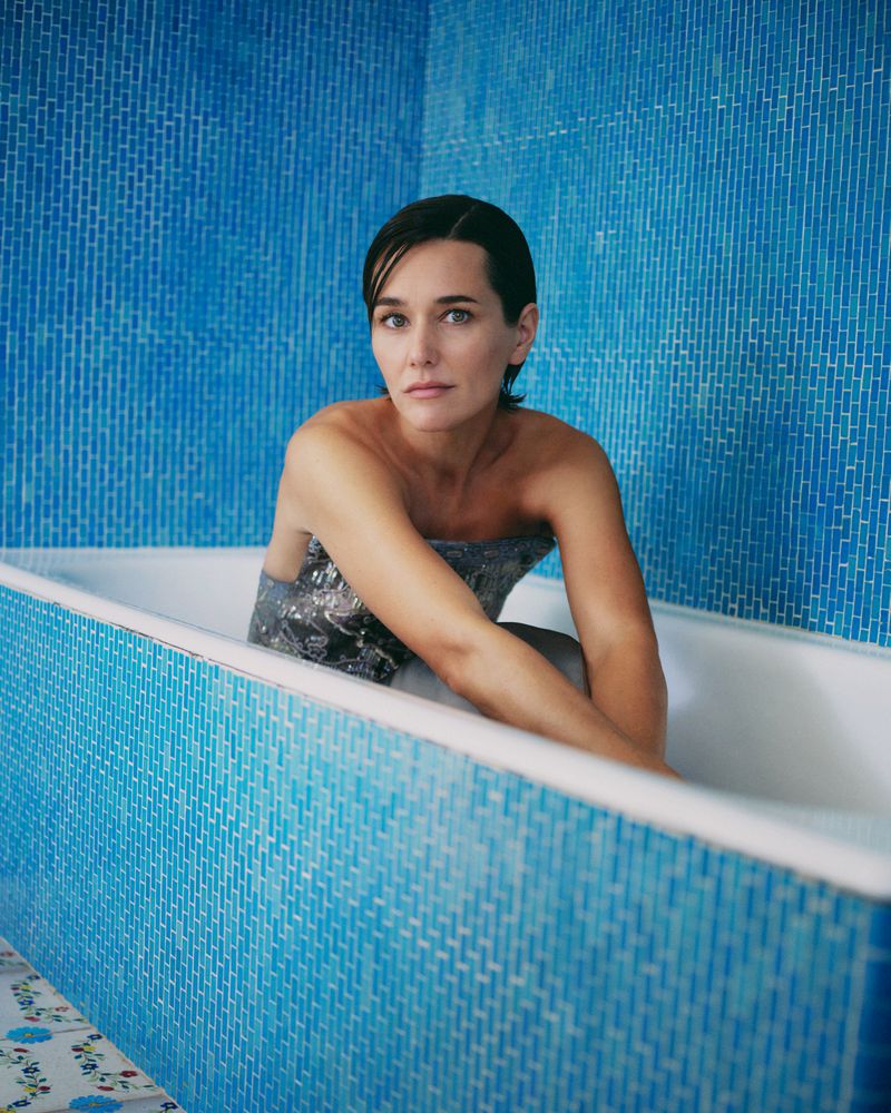 Woman with short dark hair sits in a white tub filled with water. She is set against blue tile wall in an indoor bathroom setting.