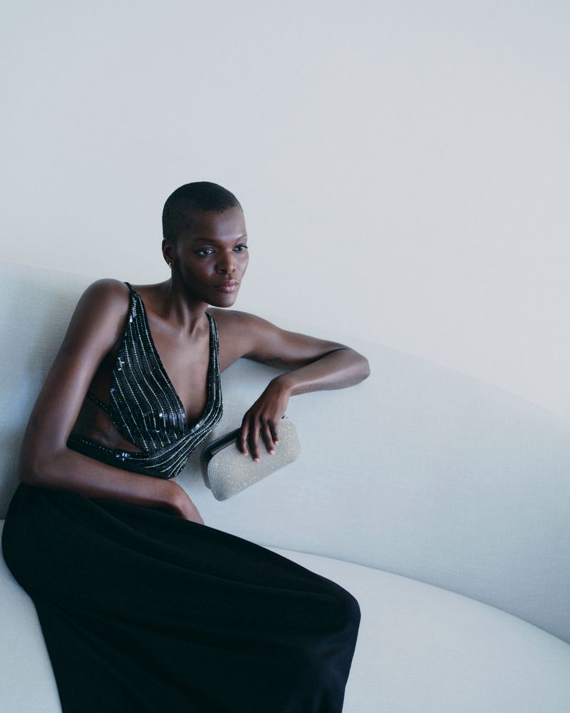 Elegant Black woman with a shaved head sits on a white couch in a cut-out black gown, holding a silver clutch in a light interior.