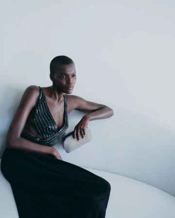 Elegant Black woman with a shaved head sits on a white couch in a cut-out black gown, holding a silver clutch in a light interior.