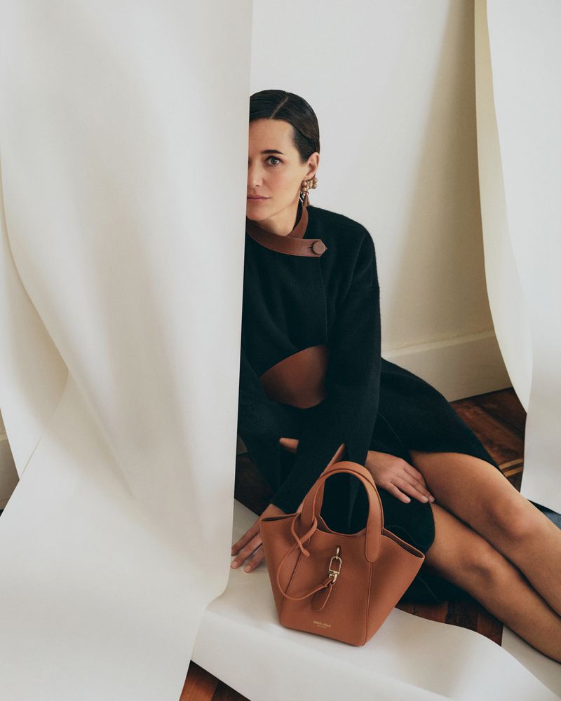 Woman in a black dress with brown leather accents sitting behind white draped paper alongside a tan leather handbag.