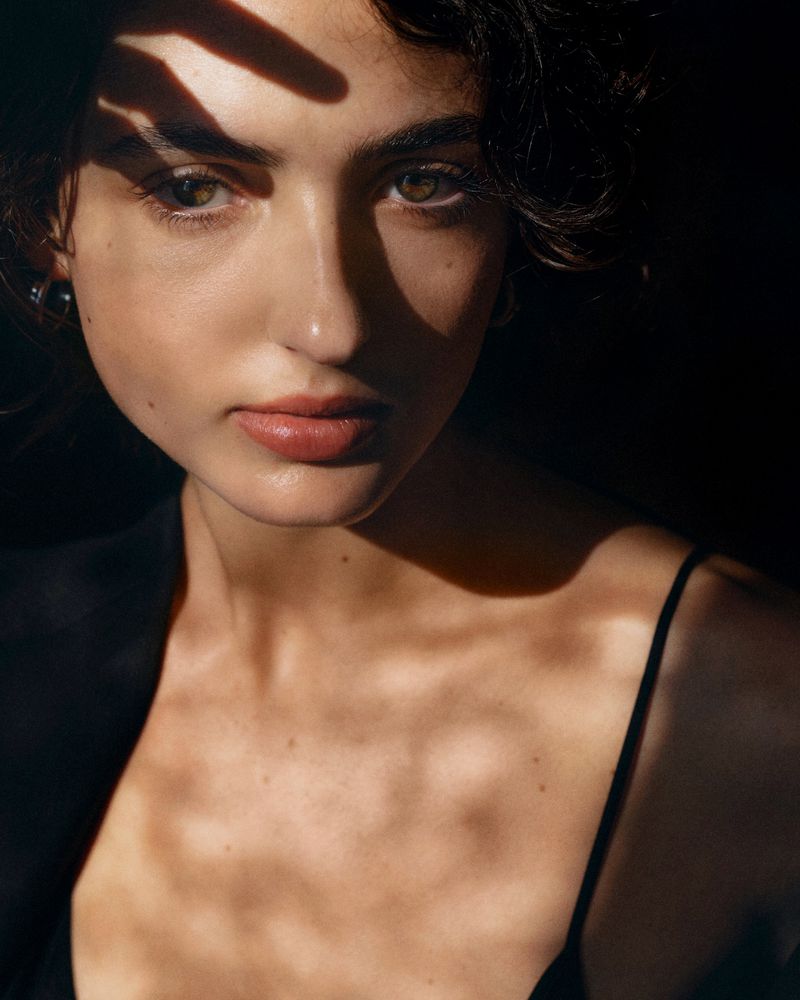 Close-up studio portrait of a young woman with dark hair and hazel eyes, wearing a black blazer, with natural light casting shadows on her face.
