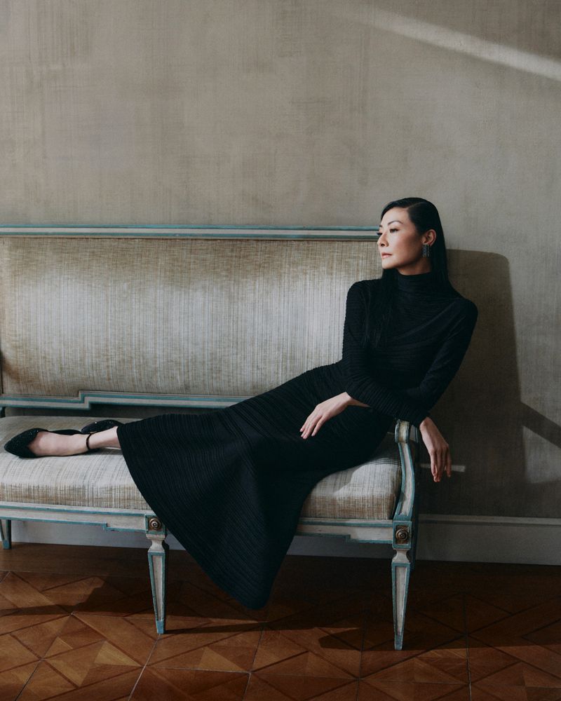 Adult female model in a black dress sits on a bench indoors with wooden floors. Light shines from the upper right.