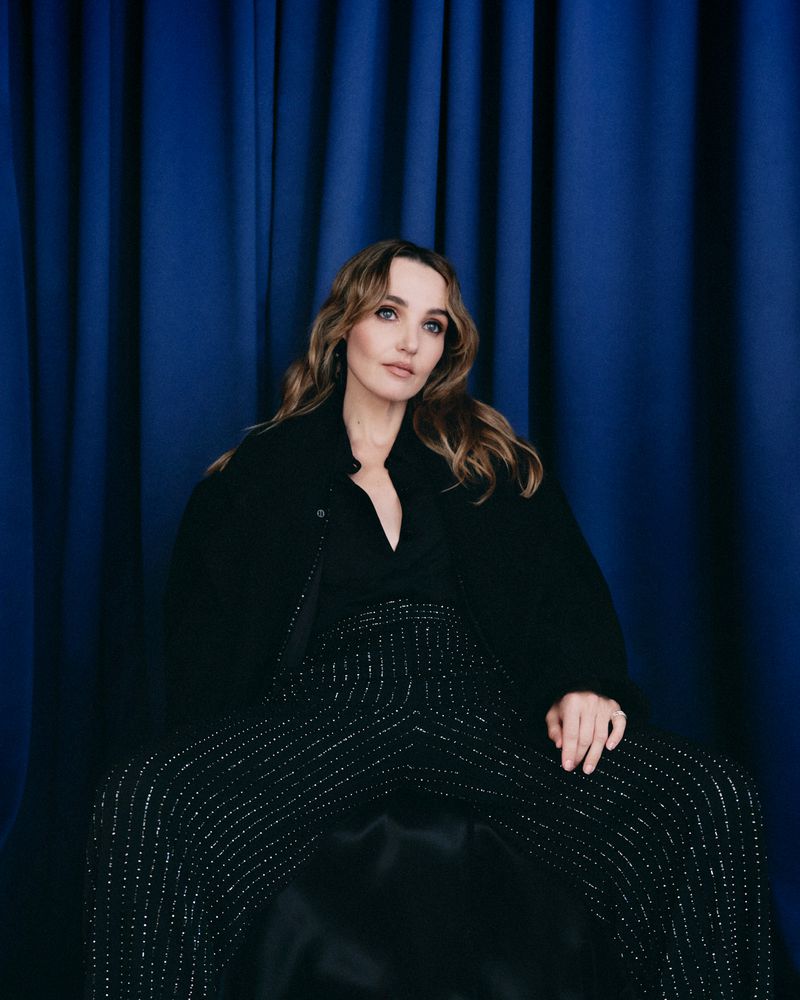 A woman with wavy, light brown hair poses in a studio with a dark blue backdrop. She wears a black jacket and vertically striped pants.