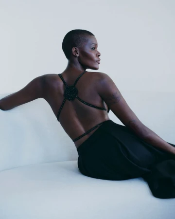 Sheila Atim wearing a black backless dress with beaded straps seated on a white sofa for Giorgio Armani.