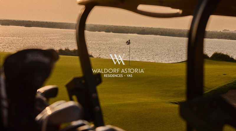 Golf course view from a golf cart with Waldorf Astoria Residences logo, featuring a green lawn, a lake, and a flag on the golf course.