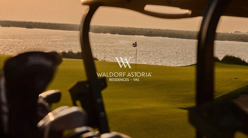 Golf course view from a golf cart with Waldorf Astoria Residences logo, featuring a green lawn, a lake, and a flag on the golf course.